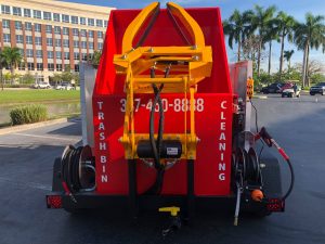 Rear view of a red trash bin cleaning truck with yellow hydraulic equipment and the phone number 337-450-8888 displayed on the back.