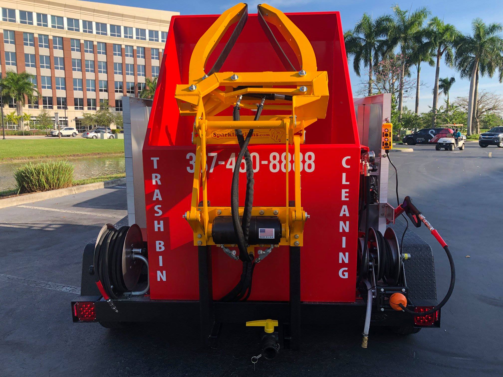 Rear view of a red trash bin cleaning truck with yellow hydraulic equipment and the phone number 337-450-8888 displayed on the back.