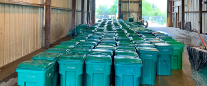 Numerous green recycling bins with closed lids are arranged in neatly defined rows inside a large warehouse.